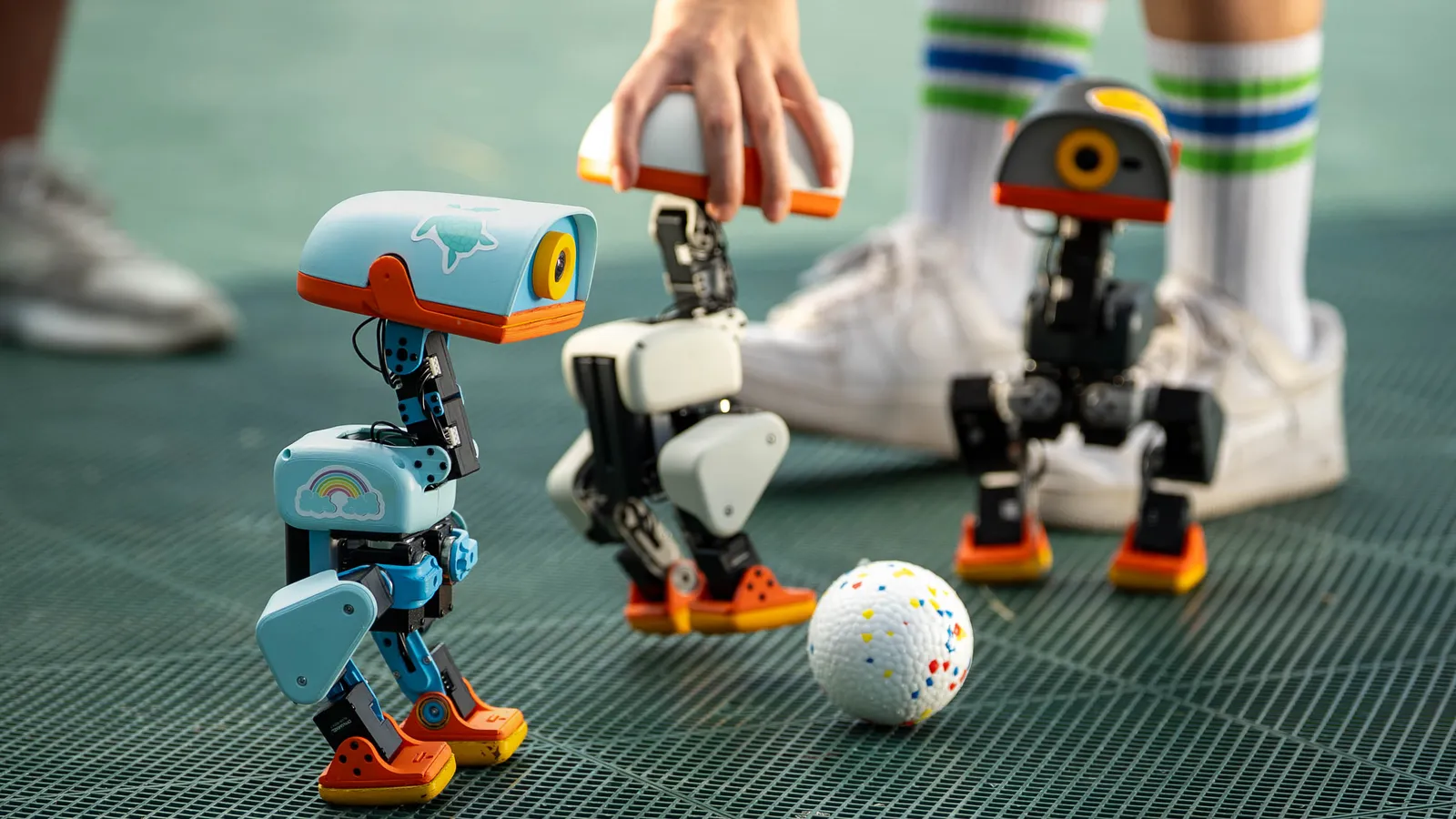 Three Microduck bipeds on a green playing surface beside a white ball, with a hand lifting one of them.